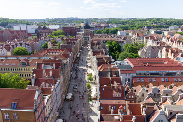 Aerial panoramic view of the historic Old Town of Gdańsk, Poland, with red tiled rooftops, bustling pedestrian street, and surrounding green landscape on a sunny summer day