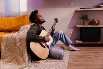 Male musician playing guitar and sitting on a blanket and leaning on a sofa