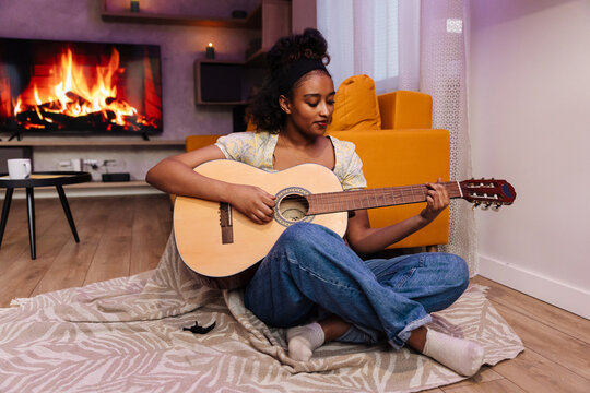 Female musician sitting on a blanket and playing the guitar and leaning on the sofa - Powered by Adobe