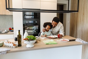 A father stands and smiles while holding a jar and hugs his daughter who is sitting at the table...