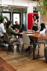 A group of four workers are sitting at a table and listening to a female worker standing next to...