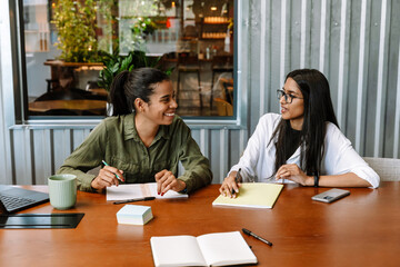 Two female workers laughing and talking while sitting at a table and holding a pencil and pen