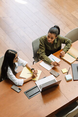 Two female workers sitting at a table and talking while holding pencils over notebooks