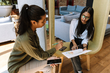A female worker holds a phone and looks at charts shown by a female worker who is talking while...