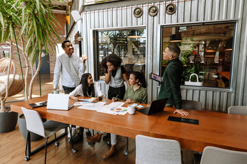 A group of three workers are talking while standing next to two female workers sitting at a table while they laugh