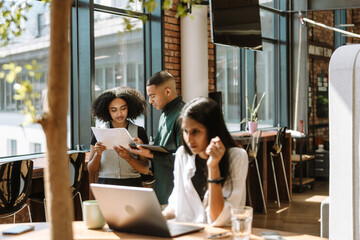 A male and female employees stand and read a document she is holding while a female employee sits next to them at a table and looks at a laptop