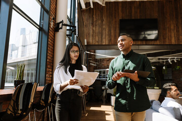 A female employee reads documents while a male employee walks and talks next to her, holding a computer tablet