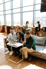 A group of three workers sit on a couch and smile while looking at a laptop where one of them is typing