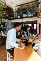 A male worker looks at a laptop that a female worker is typing on while sitting next to him at a desk while they laugh