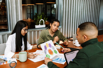 A male worker looks at a document he is holding while sitting at a desk across from two female...