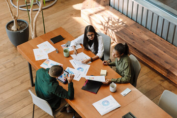 A group of three employees are sitting at a table and looking at documents that one of them is...