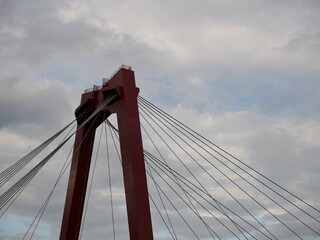 Fototapeta premium Red suspension bridge tower against dramatic cloudy sky, architectural close-up with steel cables. Engineering marvel, iconic structure, urban infrastructure, minimal color contrast.