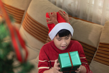 Focused young Asian boy wearing a Santa hat gently unwraps a beautifully packaged Christmas gift at home, eager to see the surprise inside.