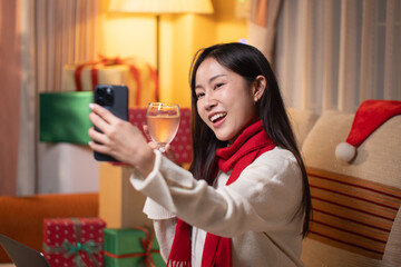 Happy young Asian woman toasting with a glass of champagne during a Christmas video call on her smartphone, celebrating the holidays with friends remotely.