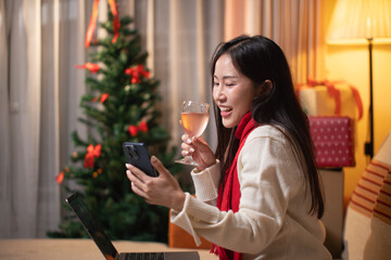 Happy young Asian woman toasting with a glass of champagne during a Christmas video call on her smartphone, celebrating the holidays with friends remotely.