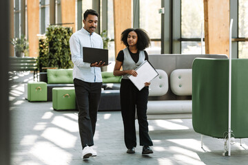 A female employee carries a folder and listens to a male employee who walks next to her and shows a...