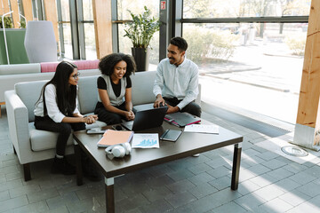 A group of three workers are laughing and sitting on a couch while talking and two of them are...