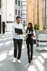 A female worker carries folders and listens to a male worker who shows her a laptop while they are...