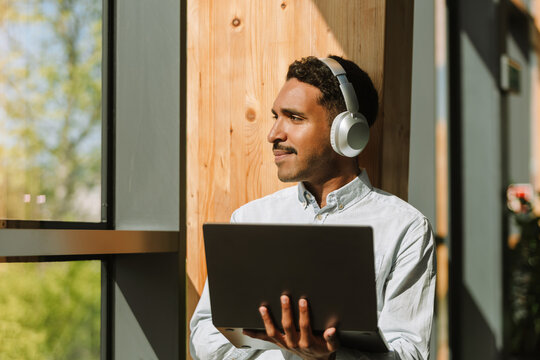 Male worker looks out the window and listens in headphones while holding a laptop and leaning on the wall