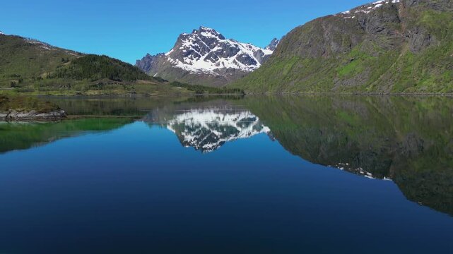Aerial view flying backwards over a calm fjord reflecting snow capped mountains in Lofoten, Norway