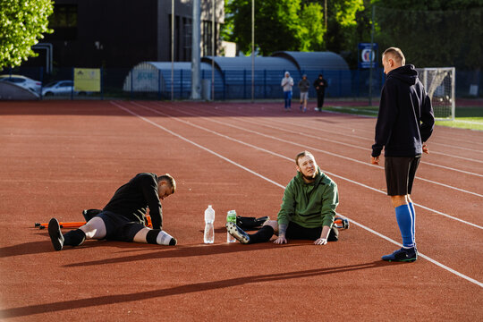 Two men sit and stretch while one of them talks to the man standing next to them