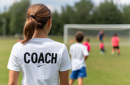 A female soccer coach watches her team practice. She is wearing a white shirt with the word 'COACH' on the back. The kids are practicing their soccer skills on a sunny day at the field.