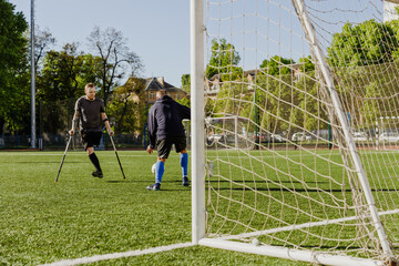 A male player on crutches watches as a male goalkeeper catches the ball while they are near the goal