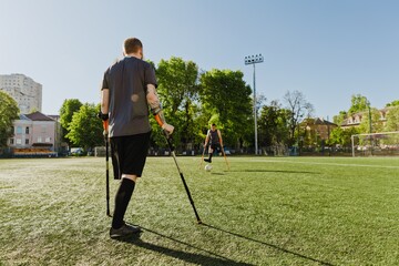 A man stands and looks at a man running towards a ball while they hold crutches