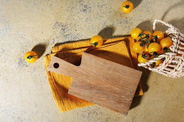 Top view of empty wooden cutting board with yellow cherry tomatoes and textured kitchen towel on rustic surface