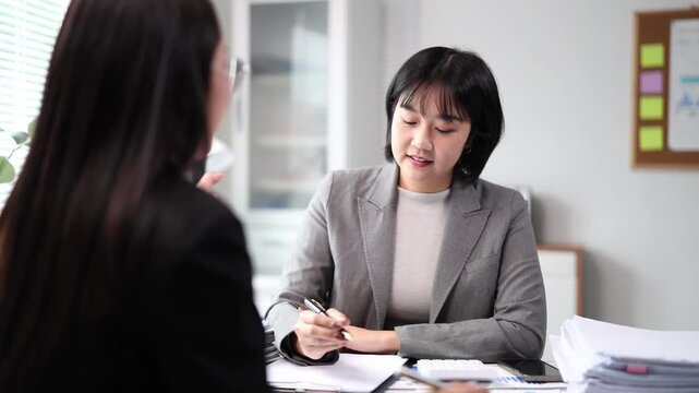 Two Asian businesswomen at an office desk review financial reports and project plans, one explaining strategy while the other listens attentively during a collaborative meeting