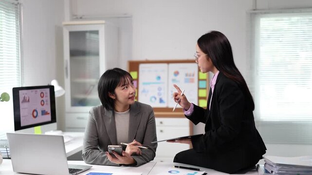 Two professional asian businesswomen collaborating on a project, analyzing charts and graphs on digital devices while having a focused discussion in a modern office environment