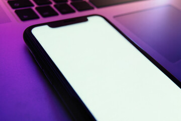 Mock up, modern smartphone with blank white screen lies on laptop keyboard under purple neon light, for app presentation design or digital background use. Space for text