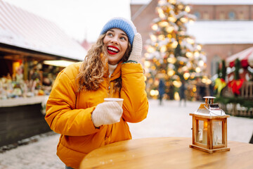 Smiling woman in yellow jacket holds hot drink at bustling winter market. Beautiful tourist enjoys New Year's atmosphere at fair decorated with lights, holding mulled wine. Concept is festive, cozy.