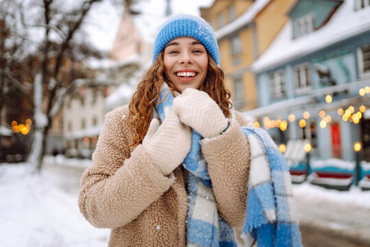 Portrait of joyful woman in cozy clothes strolling along snowy streets of city against backdrop of bright lights. A beautiful woman enjoys winter day outdoors. Concept: tourism, holiday, lifestyle.