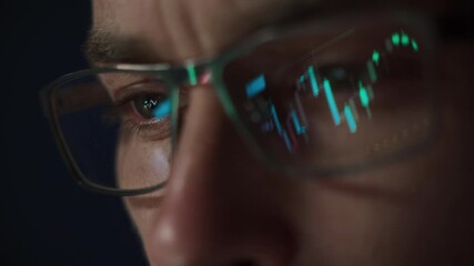Close up of focused crypto trader wearing glasses with stock market indexes reflecting in lenses while he analyzing cryptocurrency market in dark office. Trading and investment in crypto