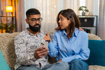 Indian couple man and woman sitting on home sofa, using smartphone for food order, joking and laughing while debating dinner. Young family enjoying playful moment with smiles and fun emotions indoors.