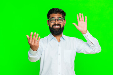 Indian man waves hand quickly with cheerful expression, inviting someone to come closer or enter with welcoming gesture. Arabian guy on green chroma key background attracting attention with smile.