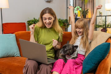 Mother and daughter with French bulldog at home finish laptop game get positive victory test result cheer clap. Woman and girl with dog on sofa celebrate win share celebrate good news smile hug reward