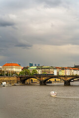 Prague Vltava River with historic stone bridge, colorful riverside buildings, baroque architecture, boats, twin spires of Vysehrad in distance, sunny