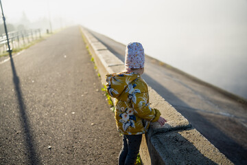Child in yellow floral jacket standing near concrete embankment overlooking foggy river on quiet...