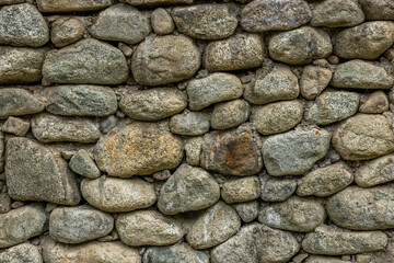 Rough stone wall constructed from many stones stands proudly in a rural outdoor setting. Daylight illuminates the textured surface, enhancing its rustic charm.