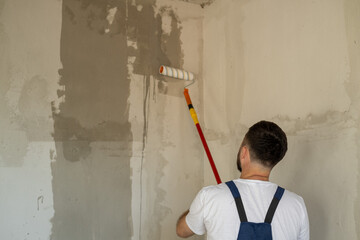 A bearded man in a work uniform is priming the walls after applying gypsum plaster.