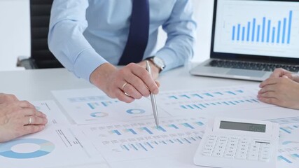 Business professional pointing at printed charts with a pen during a meeting, laptop showing bar graphs and calculator on table while team collaborates on financial analysis and planning - Powered by Adobe