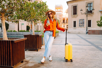 Cheerful woman with a yellow suitcase and phone admires city's architecture. A tourist in hat and...