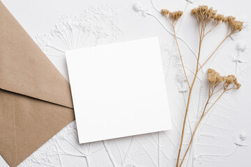 Blank square flyer card mockup with brown envelope and dried flowers on textured background, copy space for invitations, greetings or personal messages
