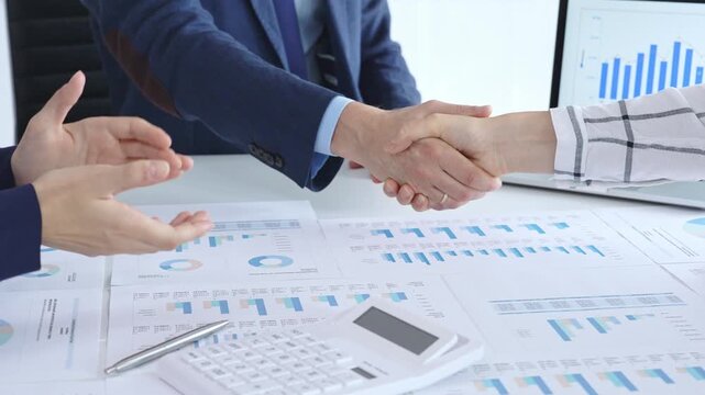 Business people are shaking hands and applauding, finalizing a deal while reviewing financial documents, graphs, calculator and charts on a desk in a modern office environment