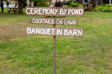 Funny rustic wooden wedding sign with white lettering directing guests to the ceremony by the pond, cocktails on the lawn, and banquet in the barn, set in an outdoor countryside venue