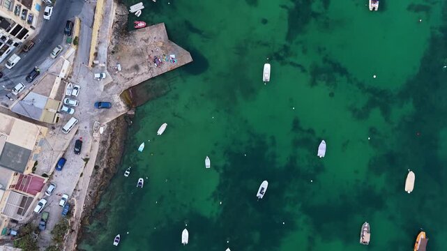4K Drone aerial of a busy coastal harbor &mdash; concrete jetty extending into clear green‑blue water, scattered small boats (anchored and drifting), adjacent urban buildings, roads, and parked vehicles