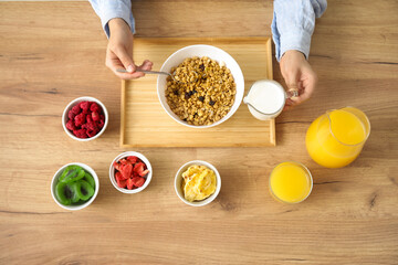 Young woman adding milk into bowl with granola on wooden table