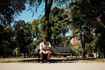 Young man in casual clothes sitting on a bench in the park and checking smartwatch. Concept of time management, technology, and relaxation